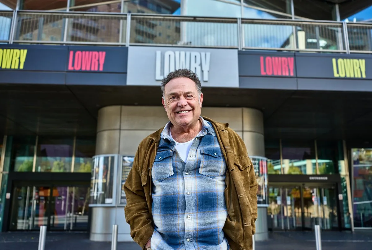 John Thomson smiling outside Lowry in Salford, wearing a tan jacket over a checked shirt