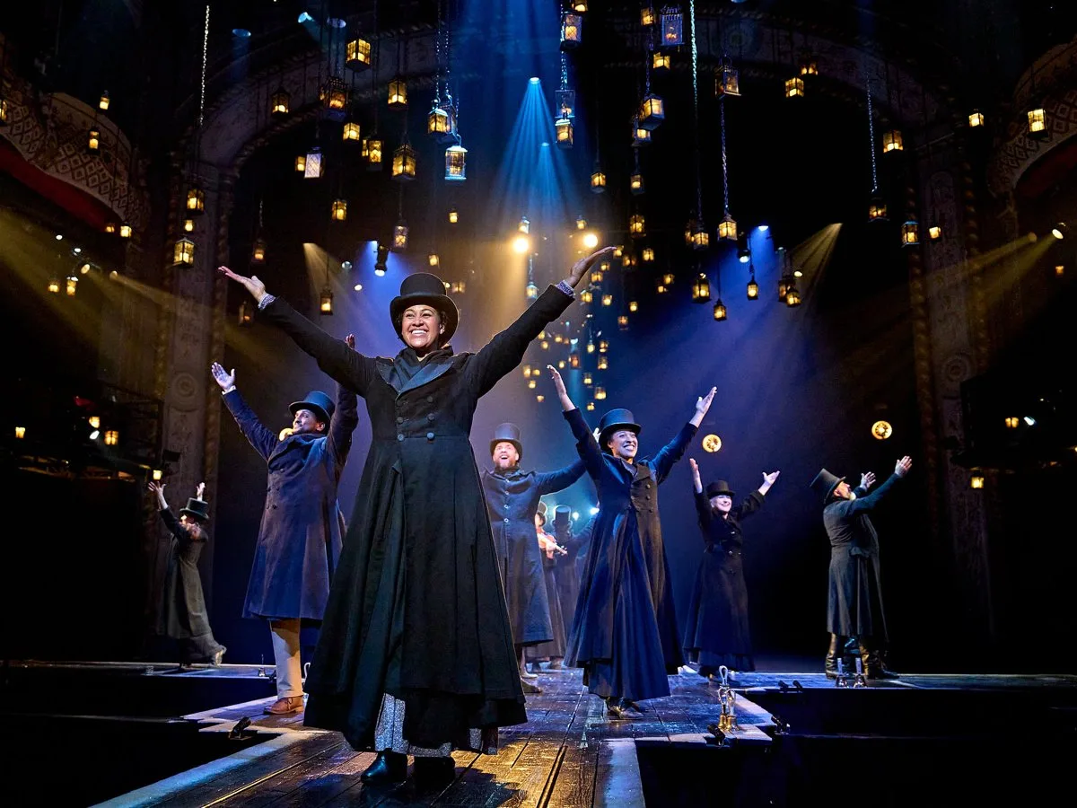 A performer in a top hat and long black coat raises their arms among the company in Victorian costume under hanging lanterns in the Old Vic production of Dickens' story