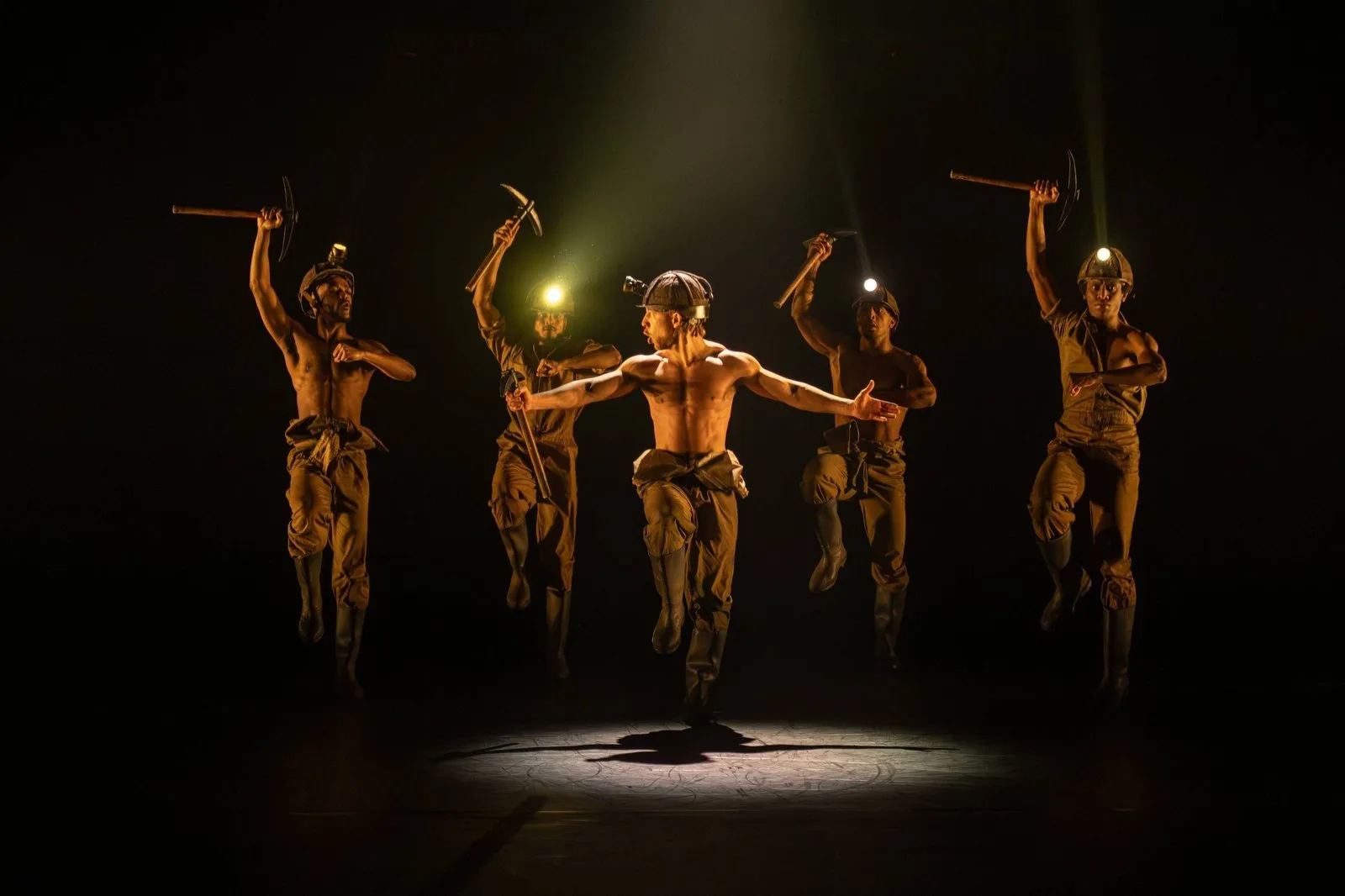 Five dancers dressed as miners leap with pickaxes raised and lamps glowing on their helmets, on a dark stage