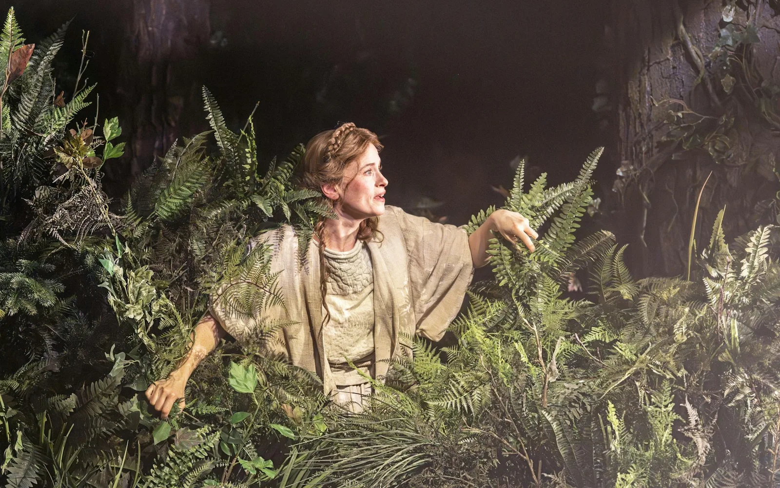 Into the Woods at the Noël Coward Theatre, with a performer peeking through ferns on the forest stage set