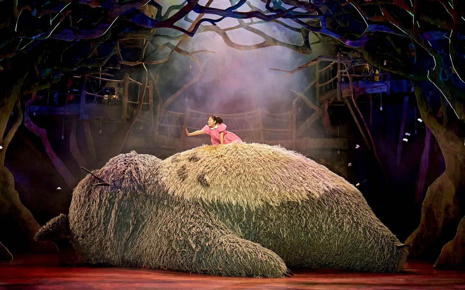 A child in a pink dress sits on the giant Totoro puppet in a misty forest at the Gillian Lynne Theatre