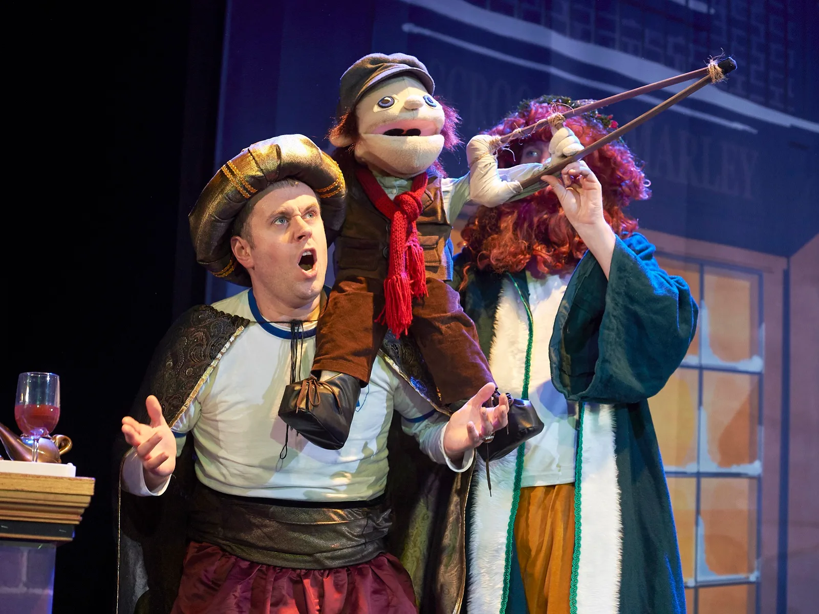 A performer in a gold turban and white tunic looks startled as a boy puppet in a flat cap rides on his shoulders, beside a performer in a crimson curly wig aiming a catapult
