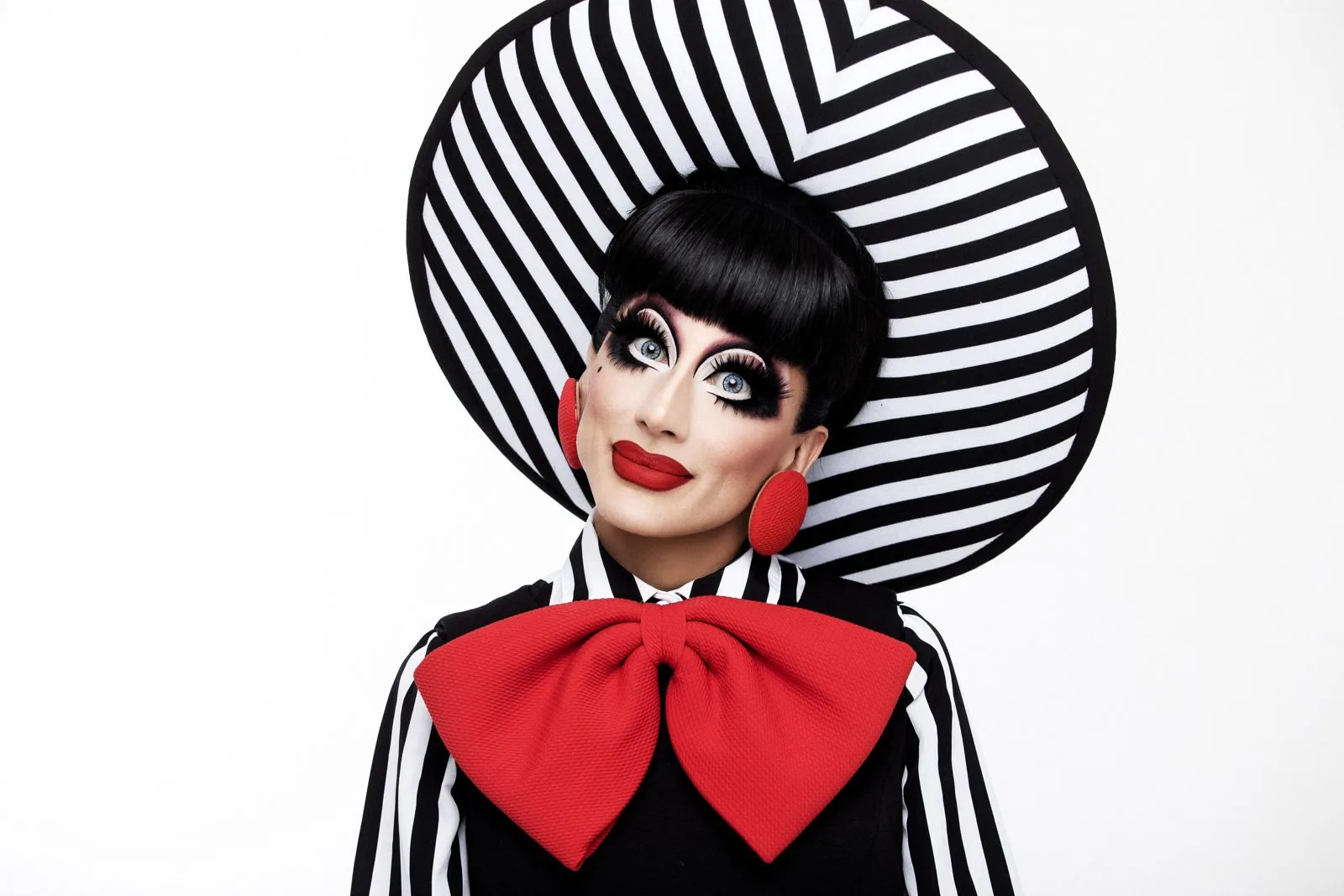Bianca Del Rio in a wide black and white striped hat, red round earrings and an oversized red bow, smiling with her head tilted against a white background