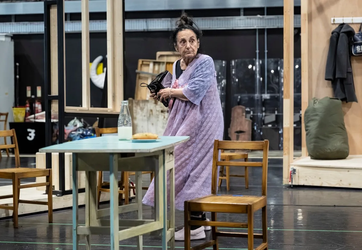 A woman with her hair pinned up, in a lilac housecoat, clutches a black handbag beside a kitchen table set with a bottle of milk and a loaf in a rehearsal room