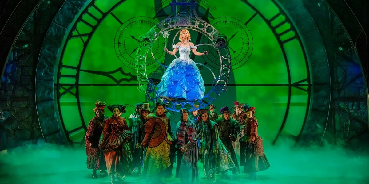 A performer in a full blue ball gown rides a silver bubble in front of a giant green clock, above a crowd of performers in colourful Oz costumes