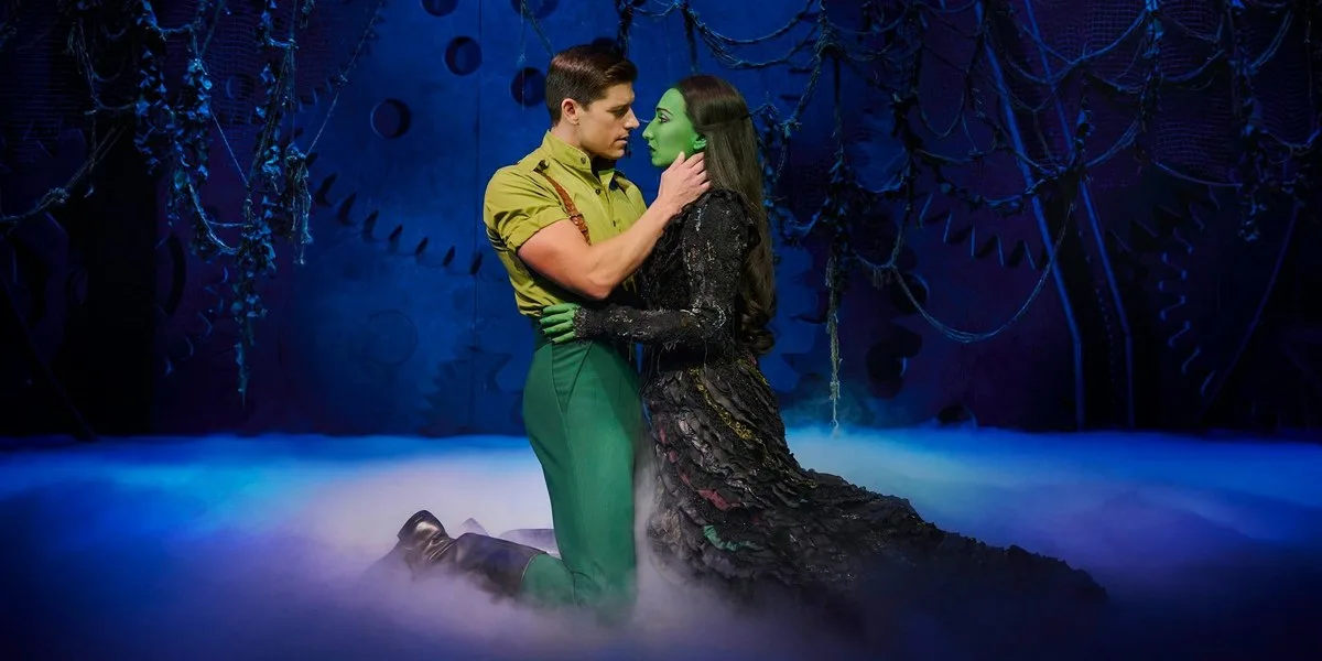 A performer in a green shirt and braces kneels in stage mist, holding a green-skinned performer in a long black dress close