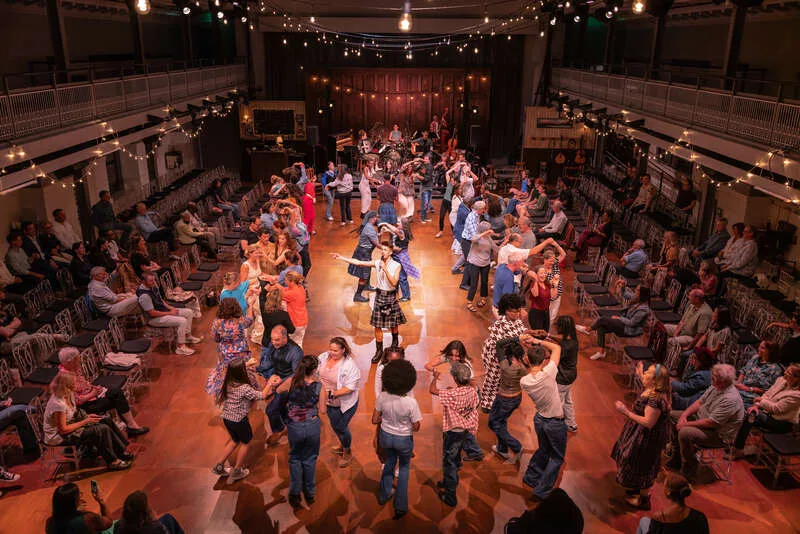 Caller in a tartan kilt directs dancers on a packed wooden floor during Ceilidh in Glasgow