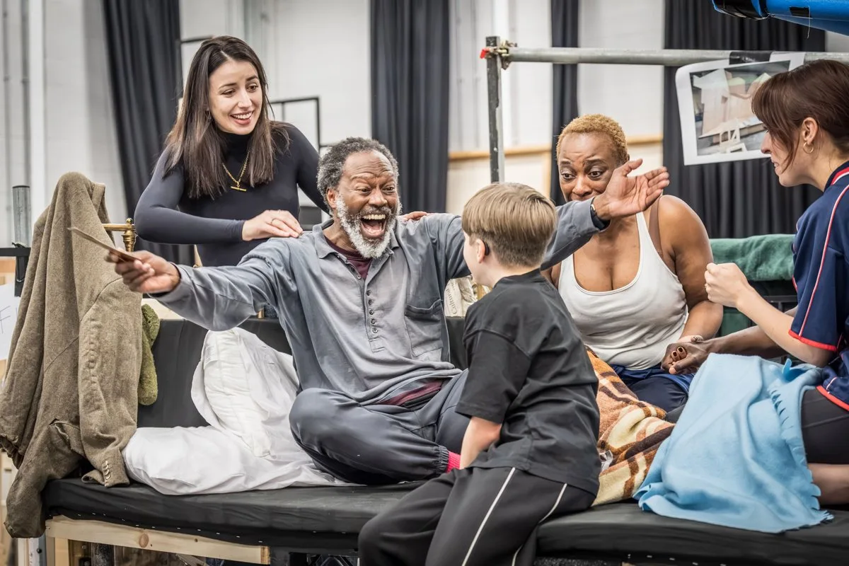 A grey bearded man sitting on a bed throws his arms wide and laughs as a young cast member playing Charlie faces him, with three women gathered around them in a rehearsal room