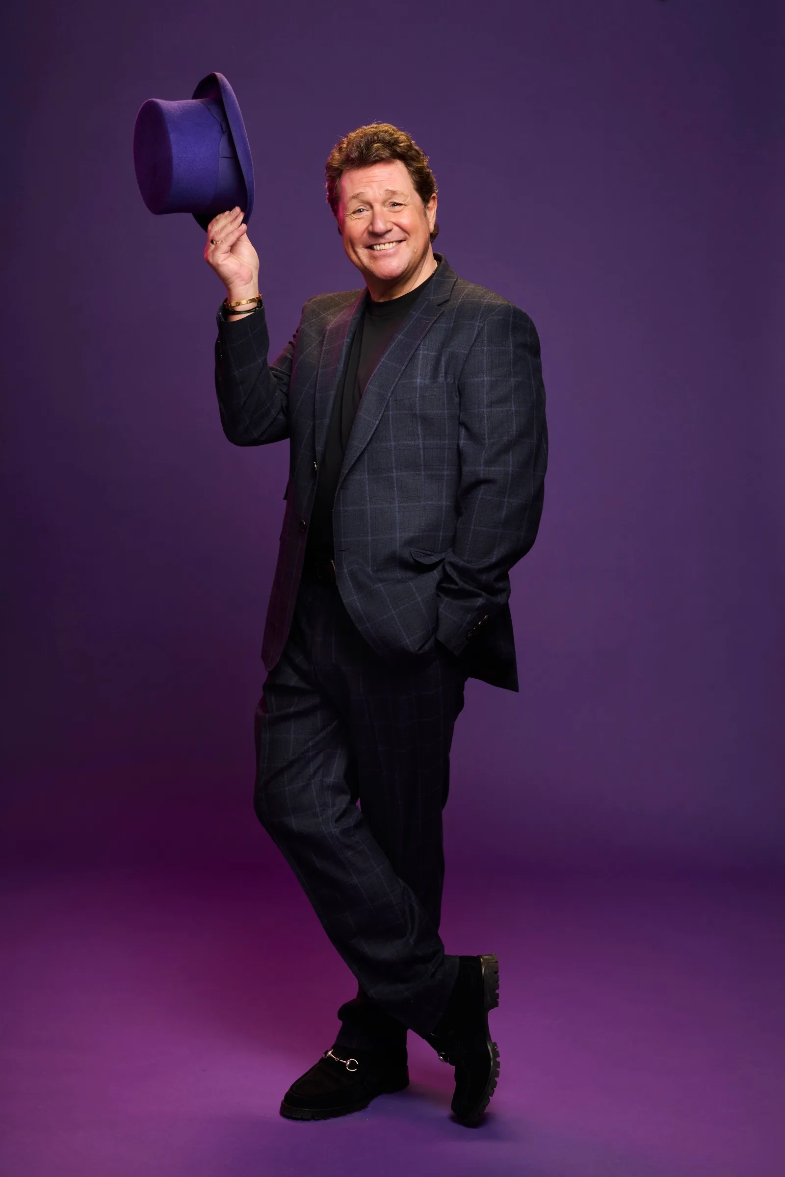A smiling performer in a dark checked suit raises a tall purple hat in front of a plum coloured studio wall
