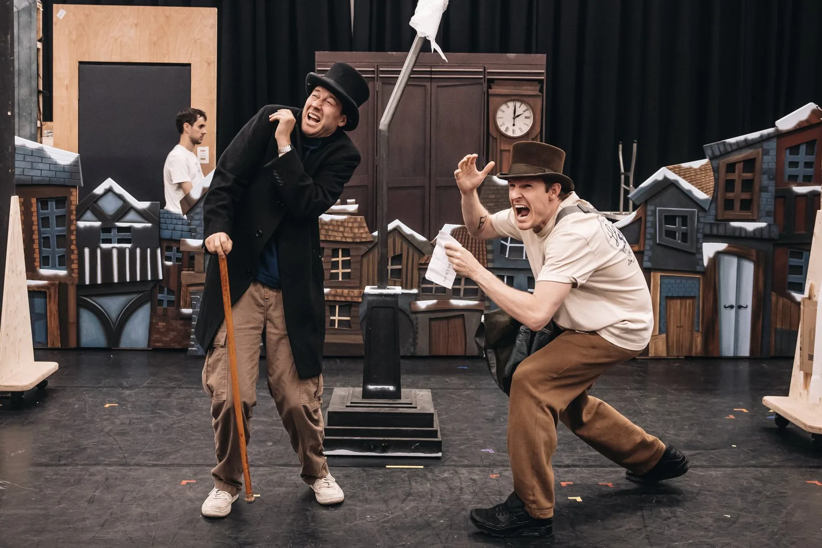 Two actors in tall dark hats lunge towards each other in a rehearsal room filled with miniature snowy rooftops and stage props