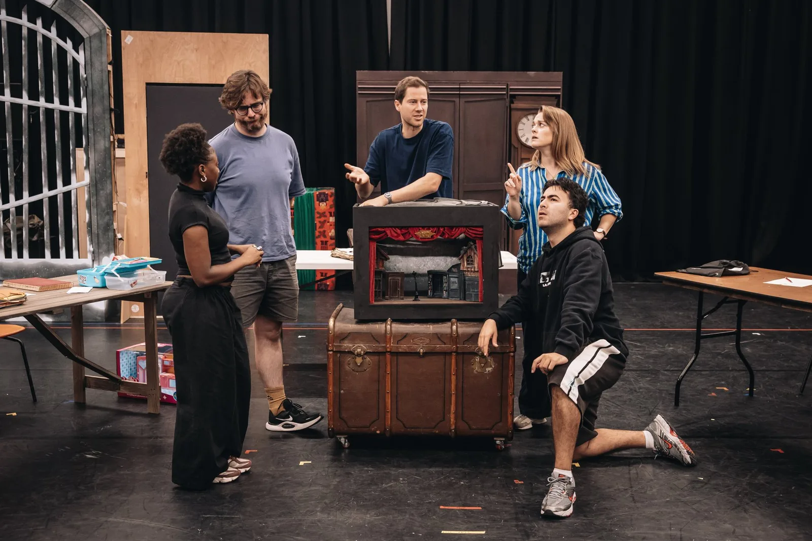 Six actors gather around a wooden trunk and a small table during a rehearsal, with painted flats stacked behind them