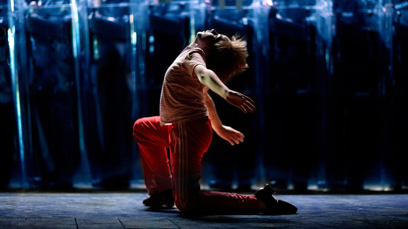 A young dancer in a crimson tracksuit kneels in front of a dark backdrop with his head thrown back and his arms flung out behind him