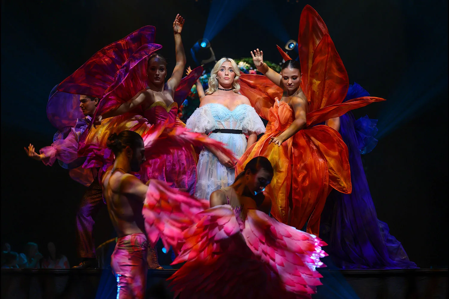 Dancers in vivid pink, orange and purple petal costumes swirl around a fair haired performer in a pale dress on a dark stage