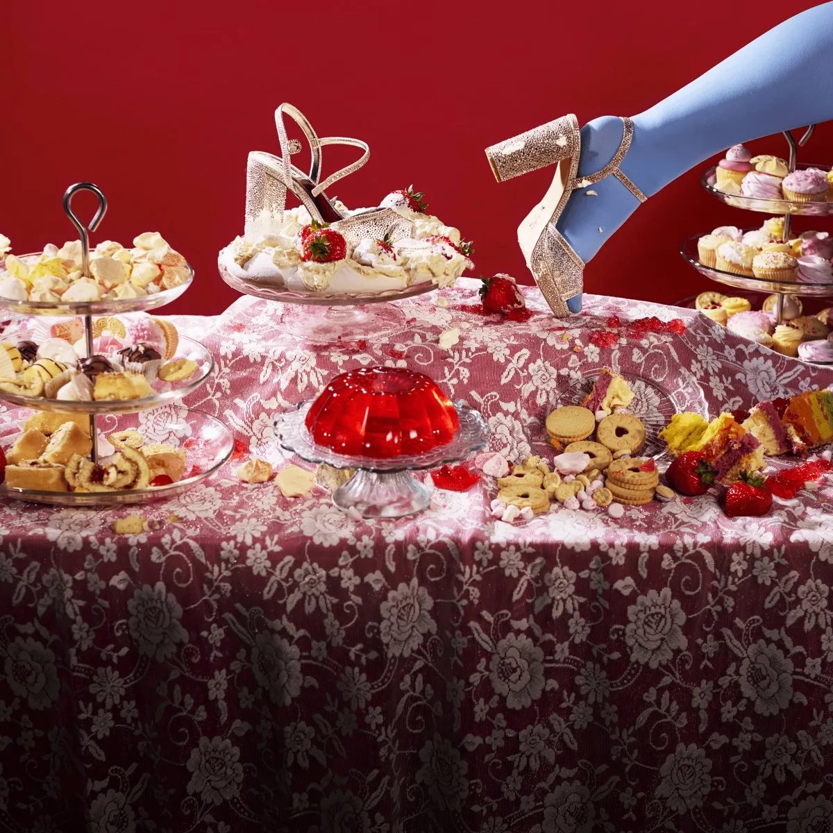 Cinderella artwork from the Citizens Theatre: a sparkly gold heeled shoe sits in a strawberry-topped pavlova on a lace-covered tea table of cakes, biscuits and a red jelly, as a foot in pale blue tights steps in from the right against a red background