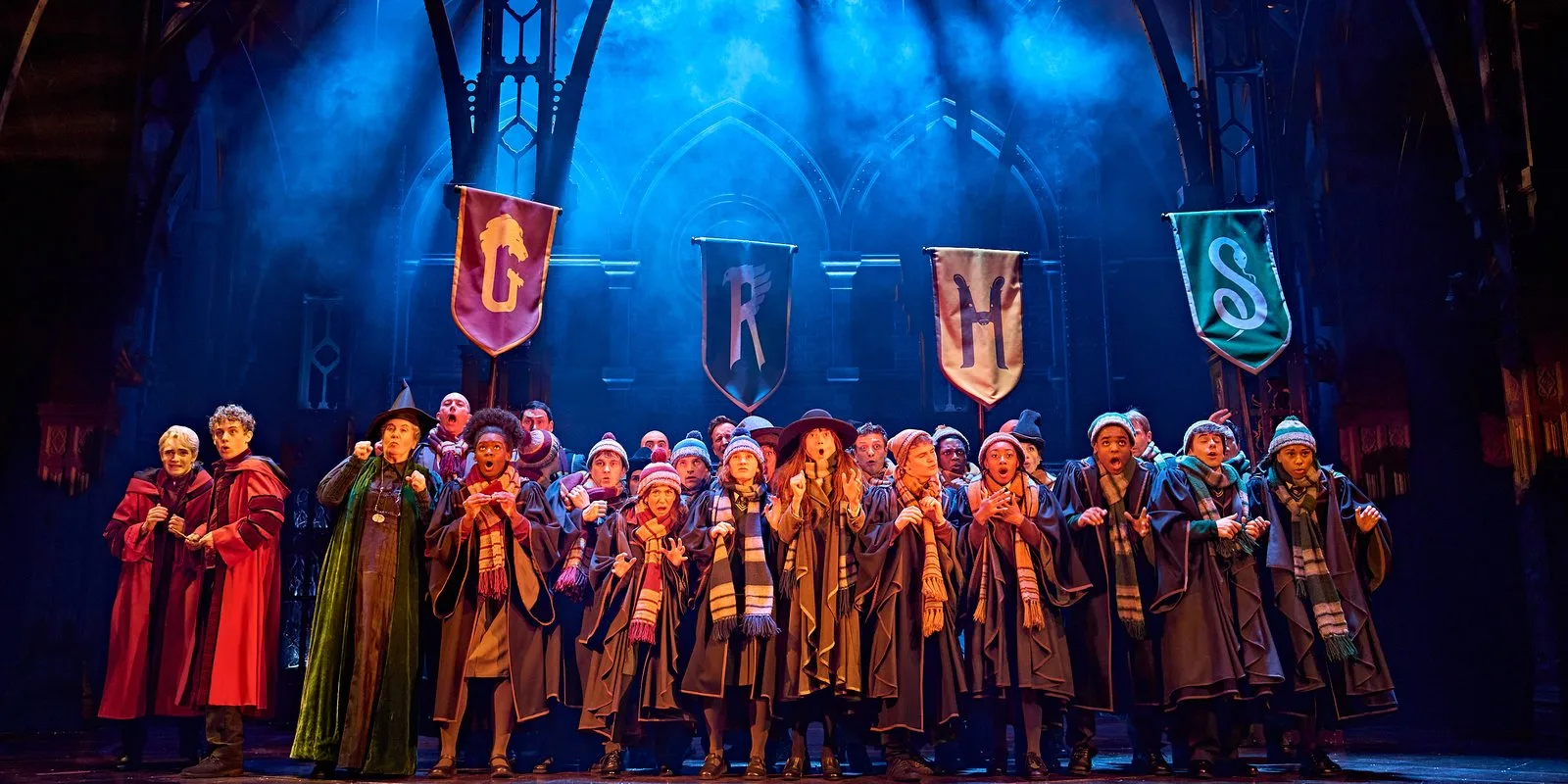 Students in Hogwarts robes and striped scarves sing together on a blue lit stage beneath four house banners