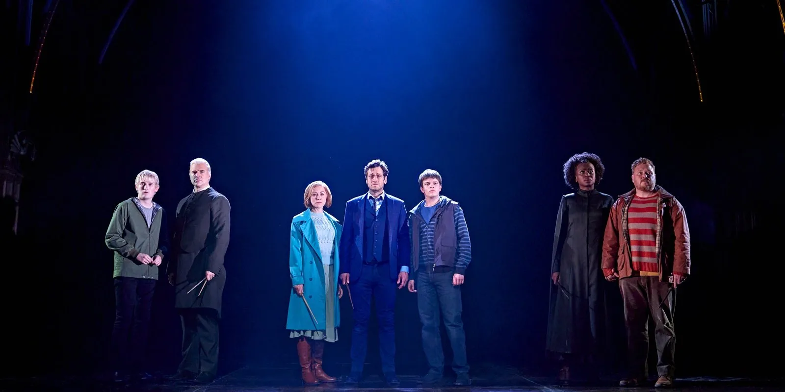 Eight actors stand in a line across a dark stage under a single shaft of blue light, some holding wands