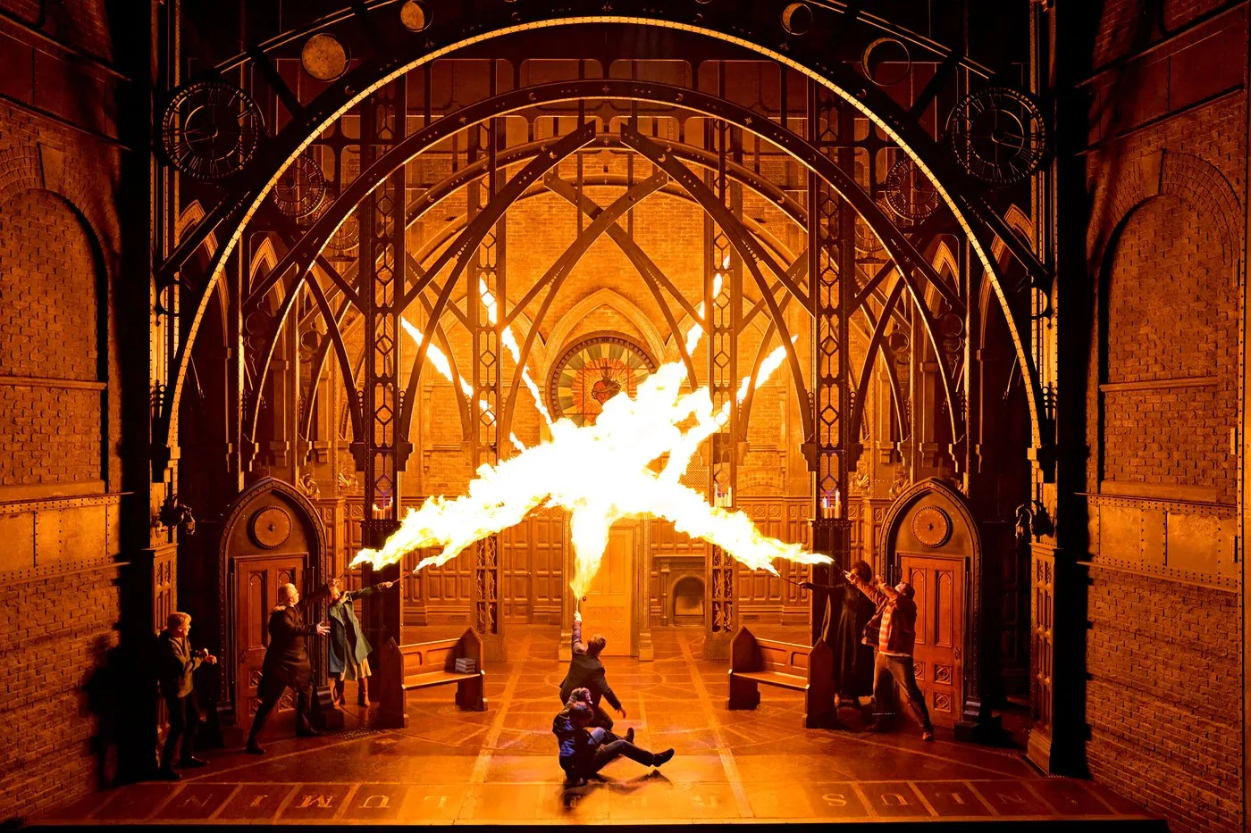 A burst of flame lights up a gothic iron archway as figures in dark robes duel on a stage glowing orange