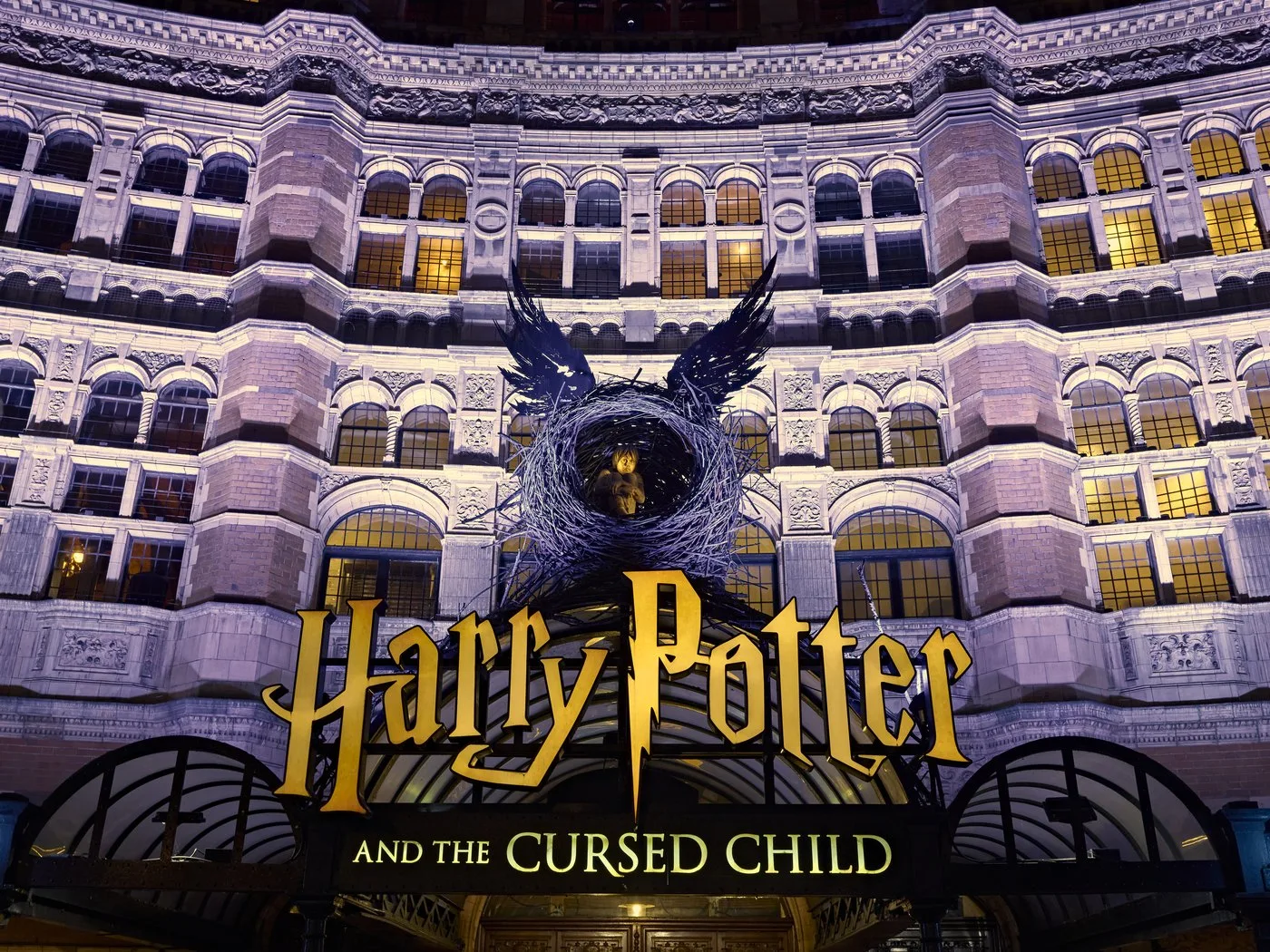 A grand brick and stone theatre facade lit up at night, with a dark winged emblem and gold lettering above the entrance