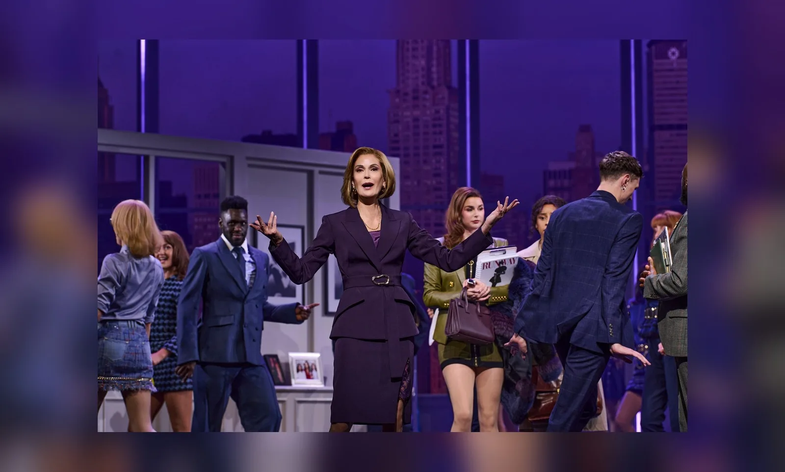 A woman in a dark fitted suit stands with her arms outstretched in a glass walled office set, surrounded by performers in smart work clothes