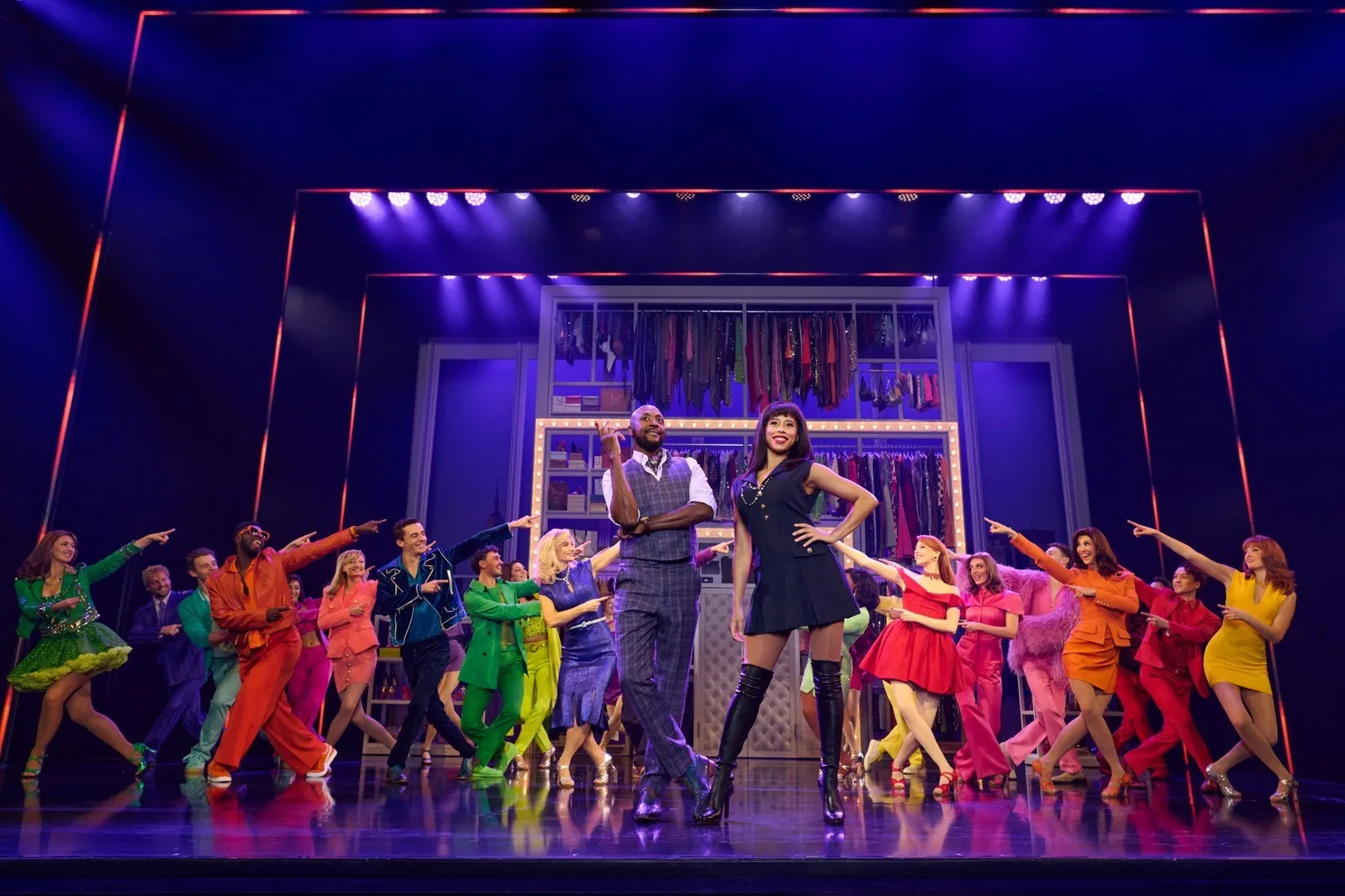 A large ensemble in brightly coloured outfits strikes poses across a stage lit in purple, with a man in a grey suit and a woman in a short dark dress at the centre