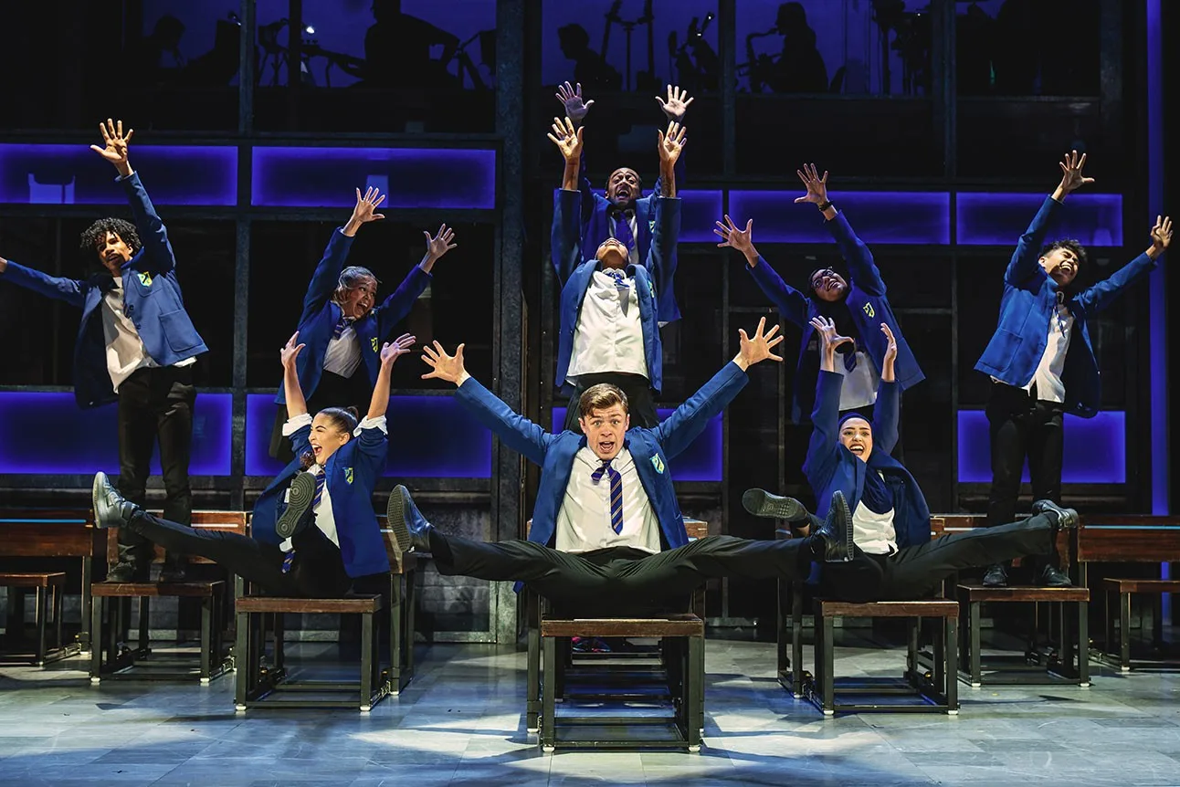 Everybody's Talking About Jamie company in Mayfield school blazers dancing on desks in a classroom musical number