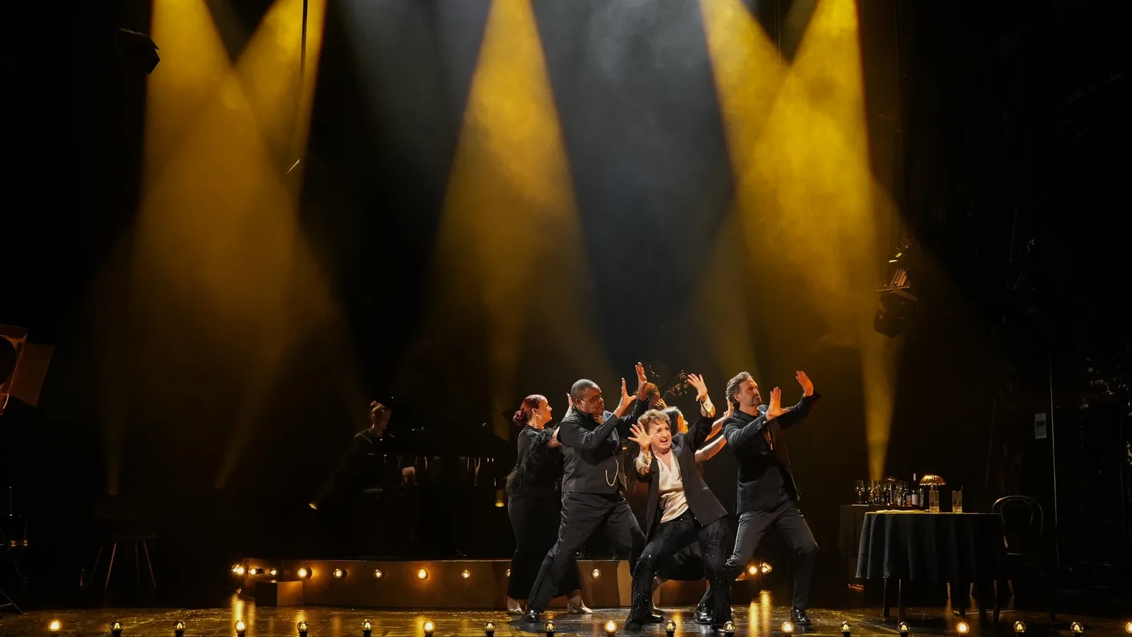 Performers in dark suits throw their arms up together beneath bright yellow spotlights as a pianist plays behind them