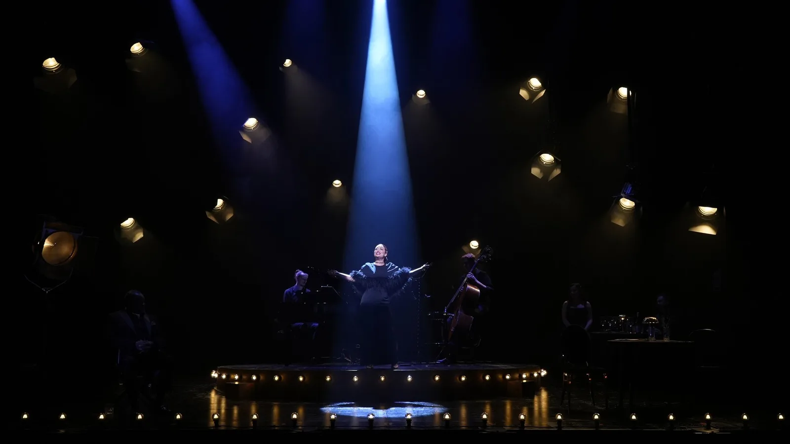 A singer in a dark feathered wrap opens her arms in a shaft of blue light, with a pianist and a cellist either side of her