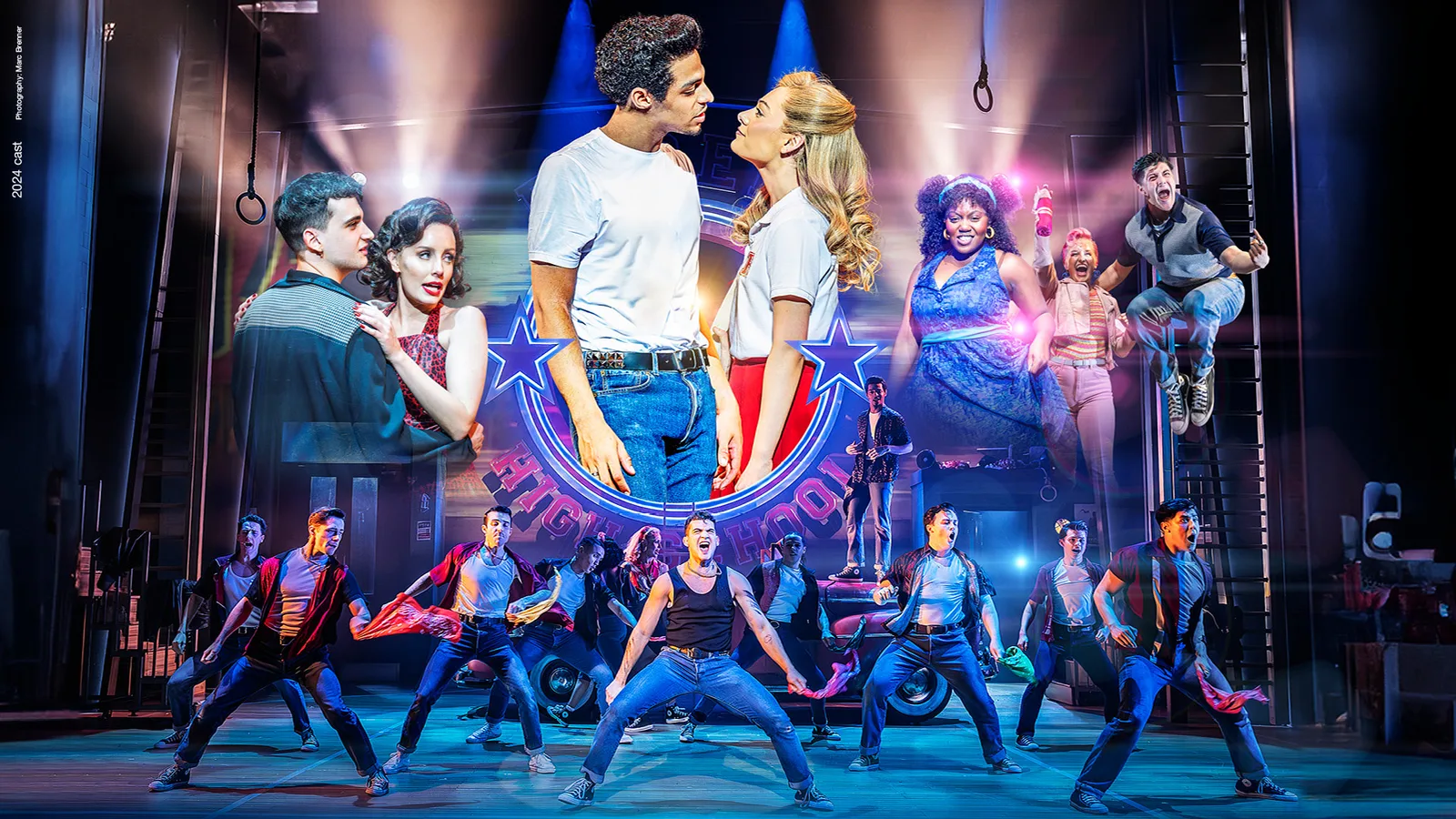 Grease UK tour image: Danny in a white T-shirt and Sandy in a white top lean in close beneath a glowing Rydell High sign, with couples either side and the ensemble dancing in denim below