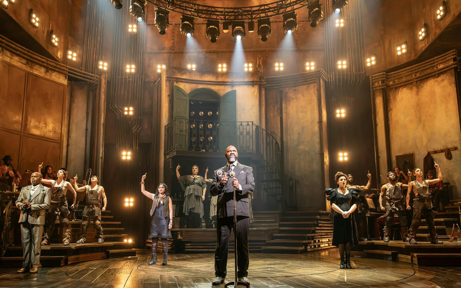 The London cast of Hadestown on stage in the West End, a performer in a pinstriped suit singing into a vintage microphone at centre as the ensemble point upwards beneath a ring of stage lights