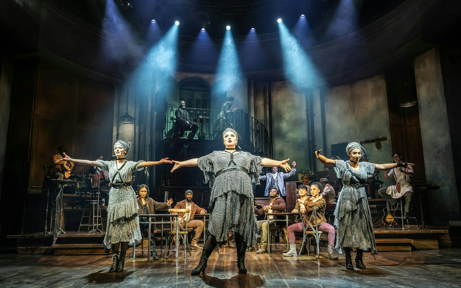 The three Fates in grey sequinned dresses and headscarves stretch their arms wide at the front of the Hadestown set, with musicians and performers seated behind them under blue spotlights
