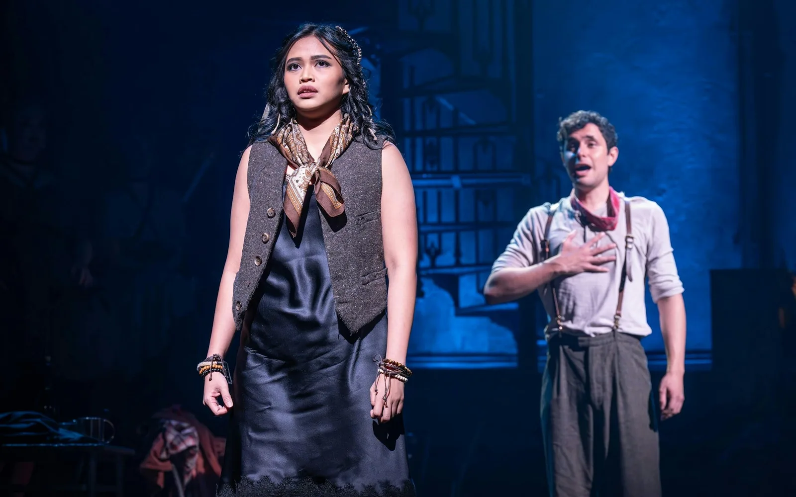Eurydice in a dark slip dress and waistcoat and Orpheus in braces and a neckerchief, lit in deep blue on the Hadestown set