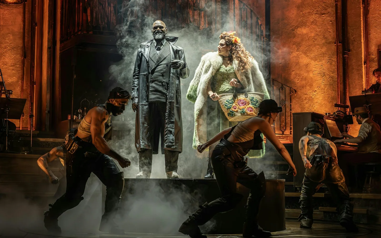 Hades in a long leather coat and Persephone in a pale fur coat stand on a raised platform in swirling haze in Hadestown, with workers crouching below