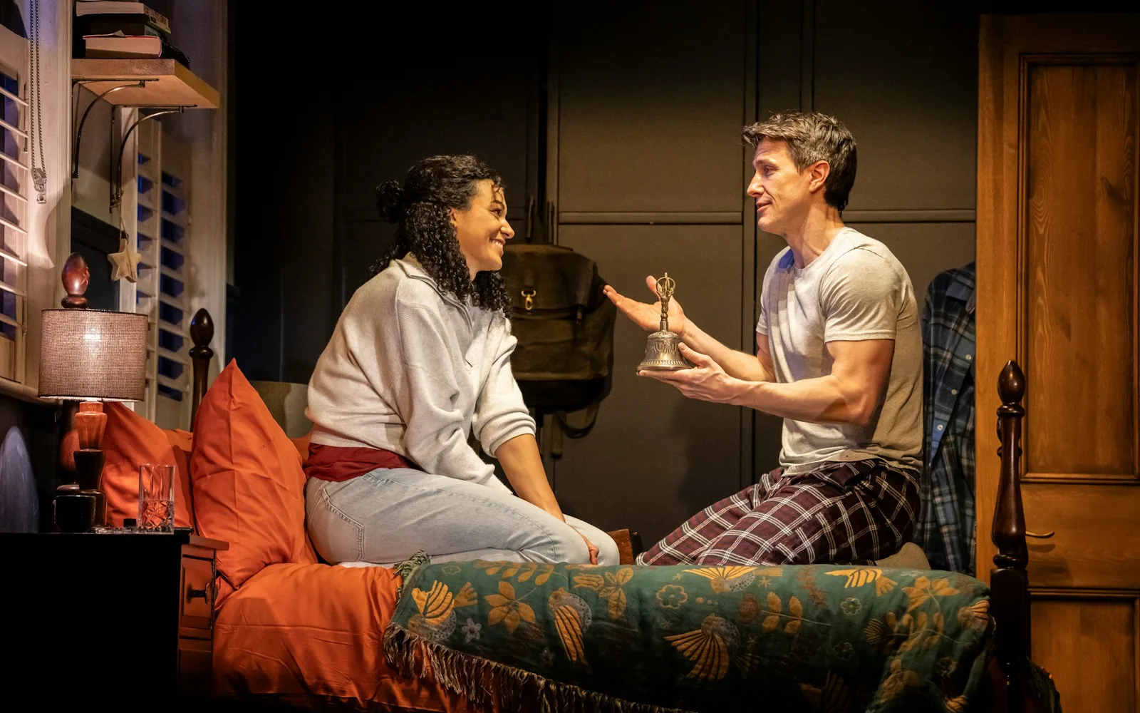 A woman in a grey top and a man in a white T-shirt and checked pyjama trousers are perched on a bed in a lamplit bedroom, the man holding up a small bell