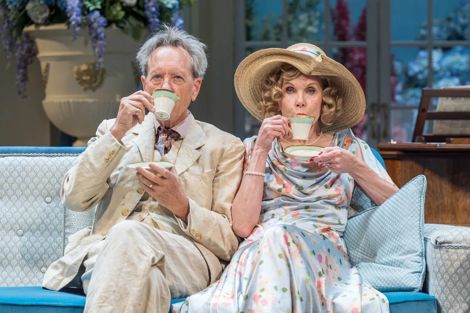 Richard E. Grant and Christine Baranski take tea on a pale blue sofa in a country house drawing room