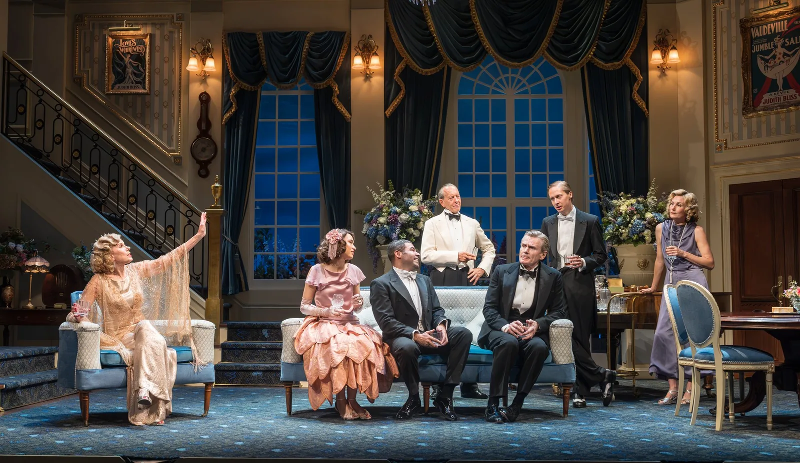 Wide view of an elegant drawing room set with a sweeping staircase, tall blue drapes and guests seated on pale sofas