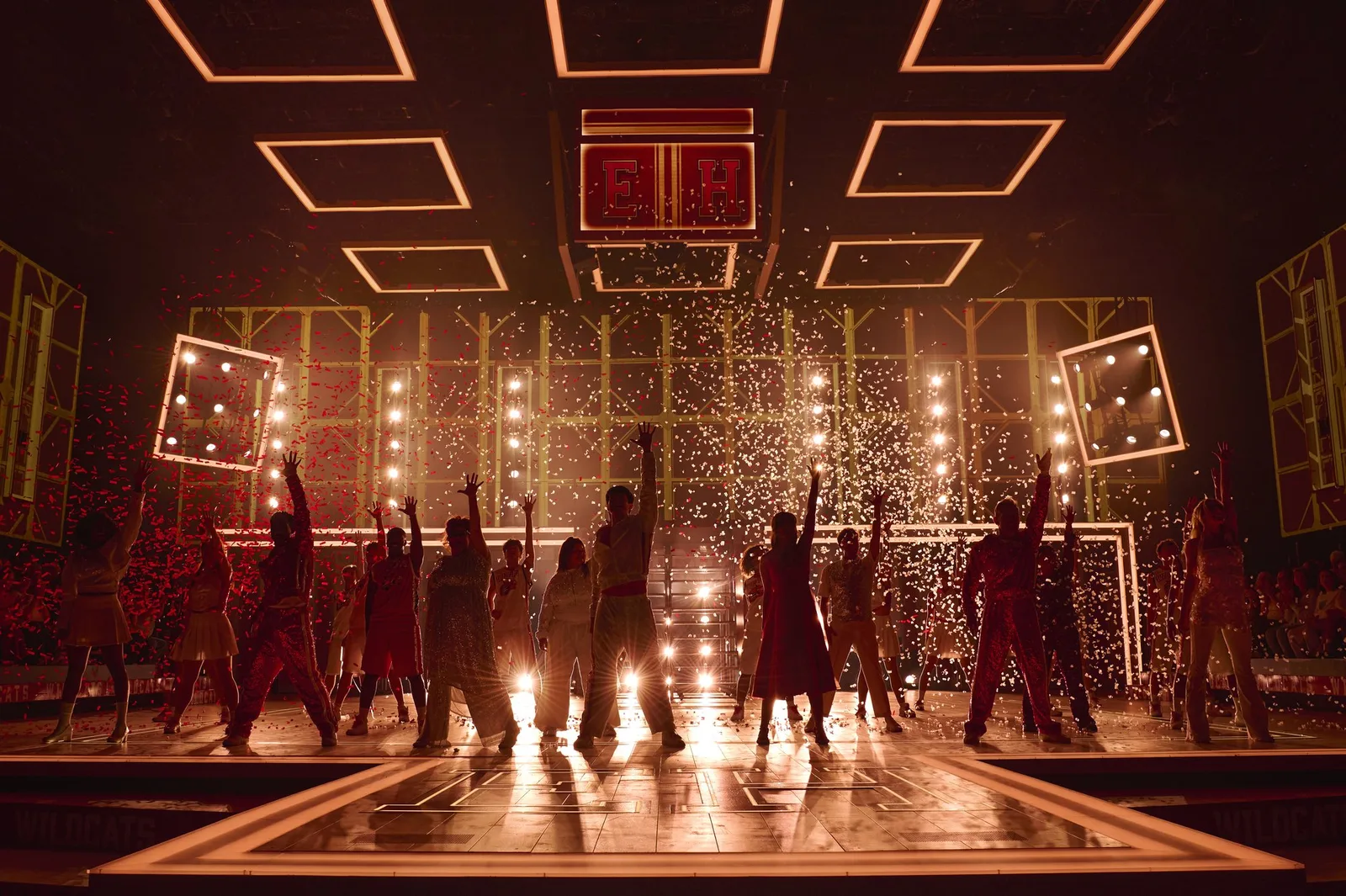 Performers raise their arms as glitter rains down in front of a wall of bright stage lights