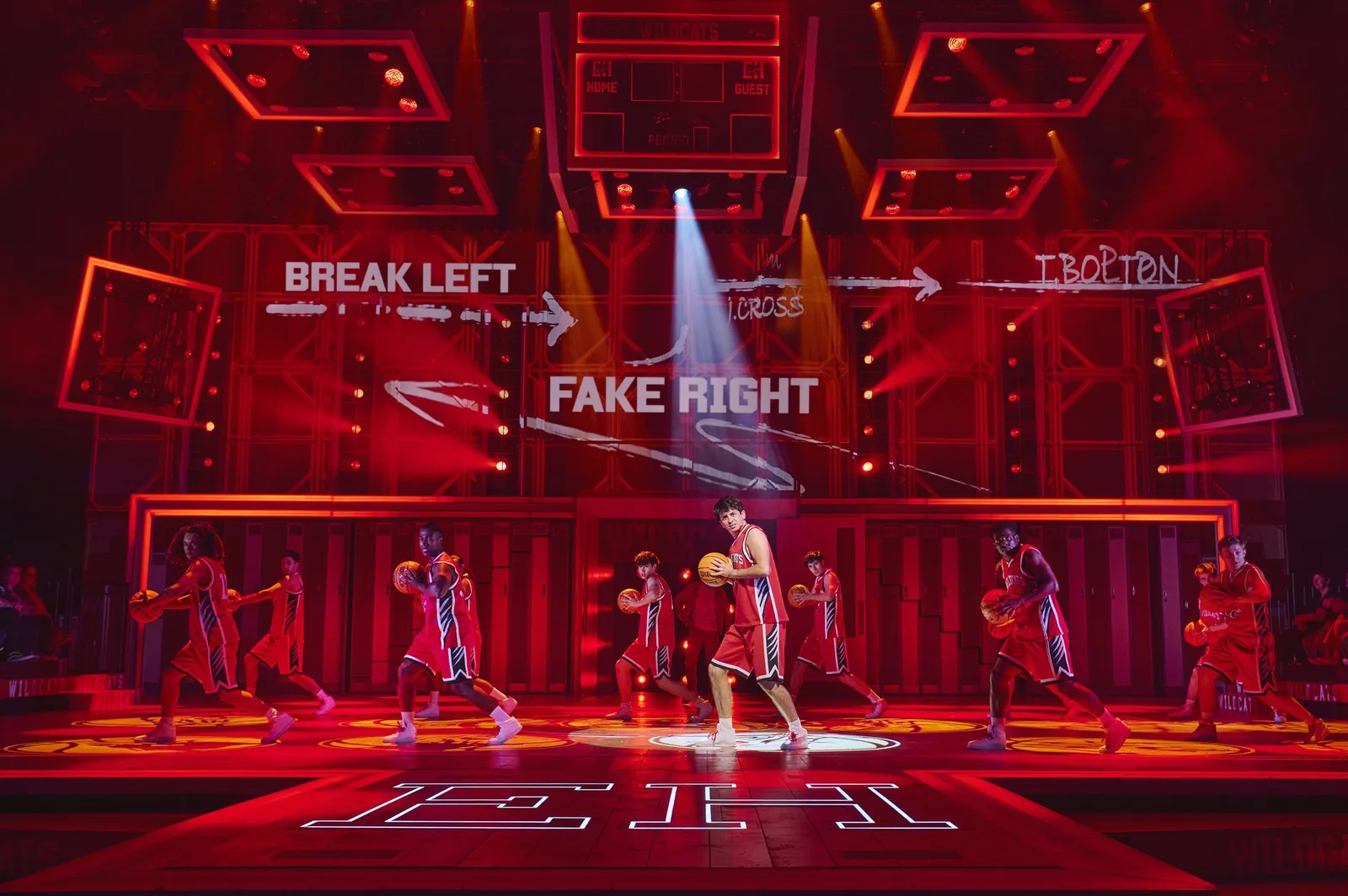Dancers in crimson and white kit leap across a basketball court under a crimson glow, with arrows on a screen behind them