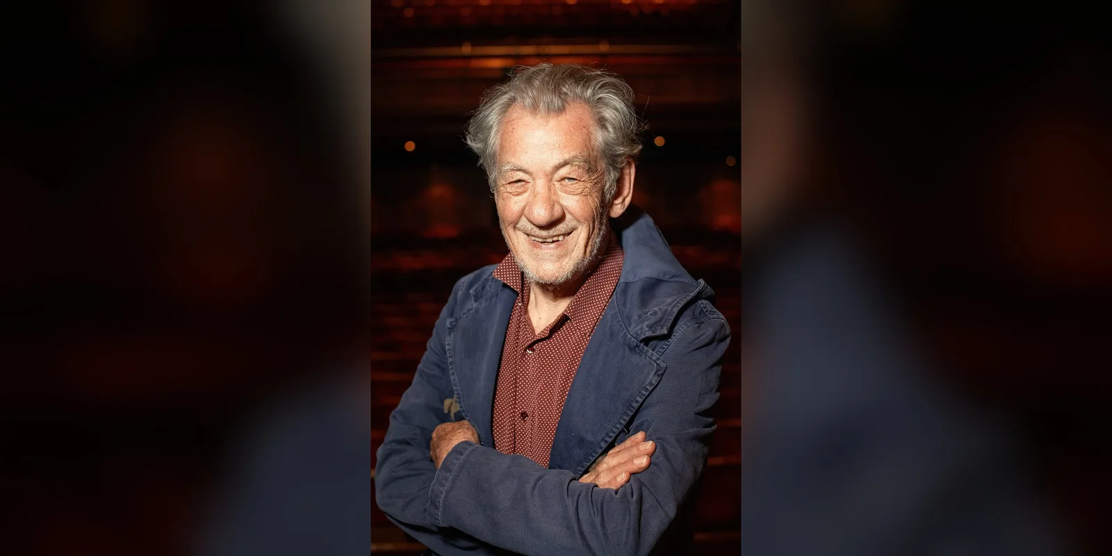 Ian McKellen smiling with his arms folded, wearing a blue jacket, in front of rows of seats in a theatre auditorium