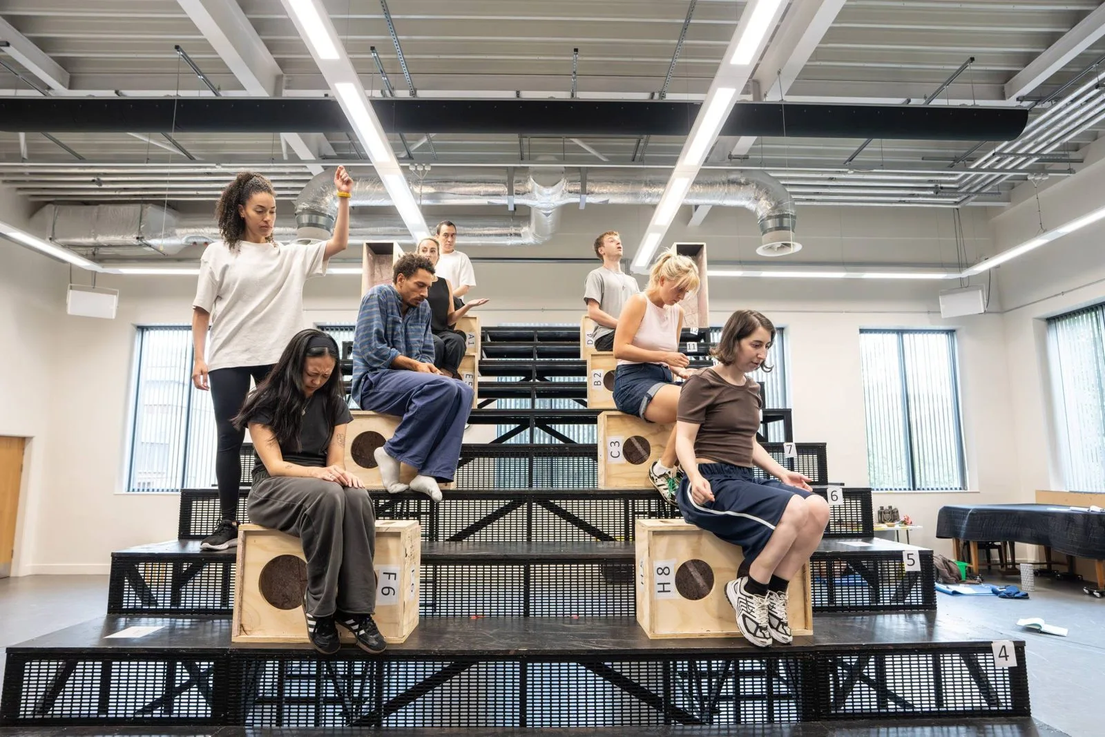 The Jane Eyre company rehearsing on tiered black steel staging with numbered wooden boxes in a bright rehearsal studio