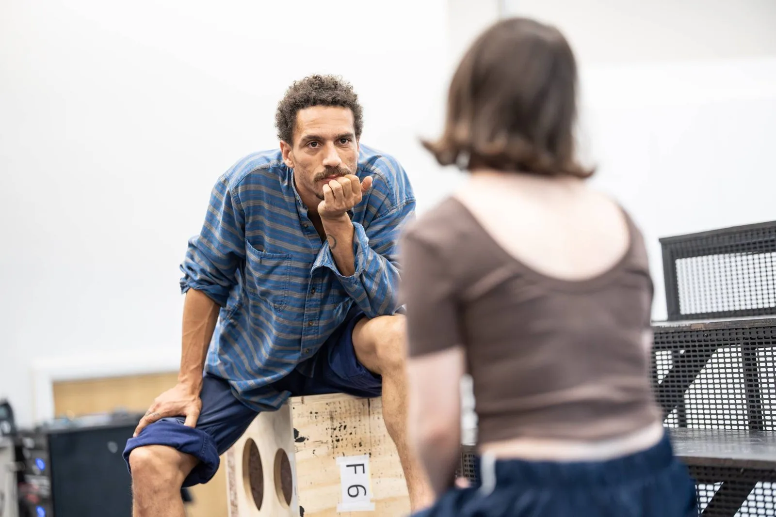 Gabriel Akuwudike in rehearsal for Jane Eyre, seated on a numbered wooden box with his chin resting on his hand as he faces a fellow actor