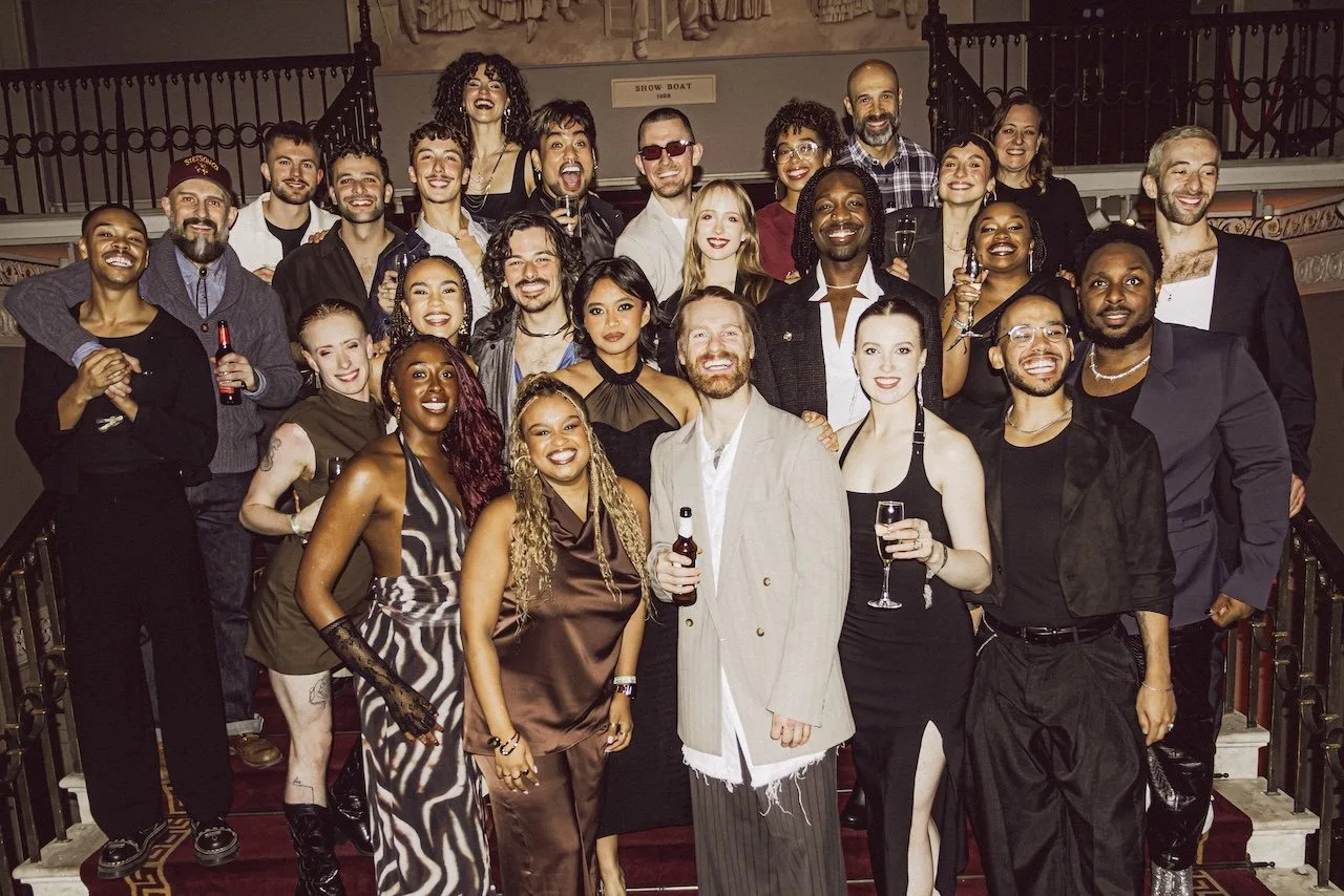 A large group of performers in evening wear pose together on a grand crimson carpeted staircase, with a bearded man in a pale suit at the centre