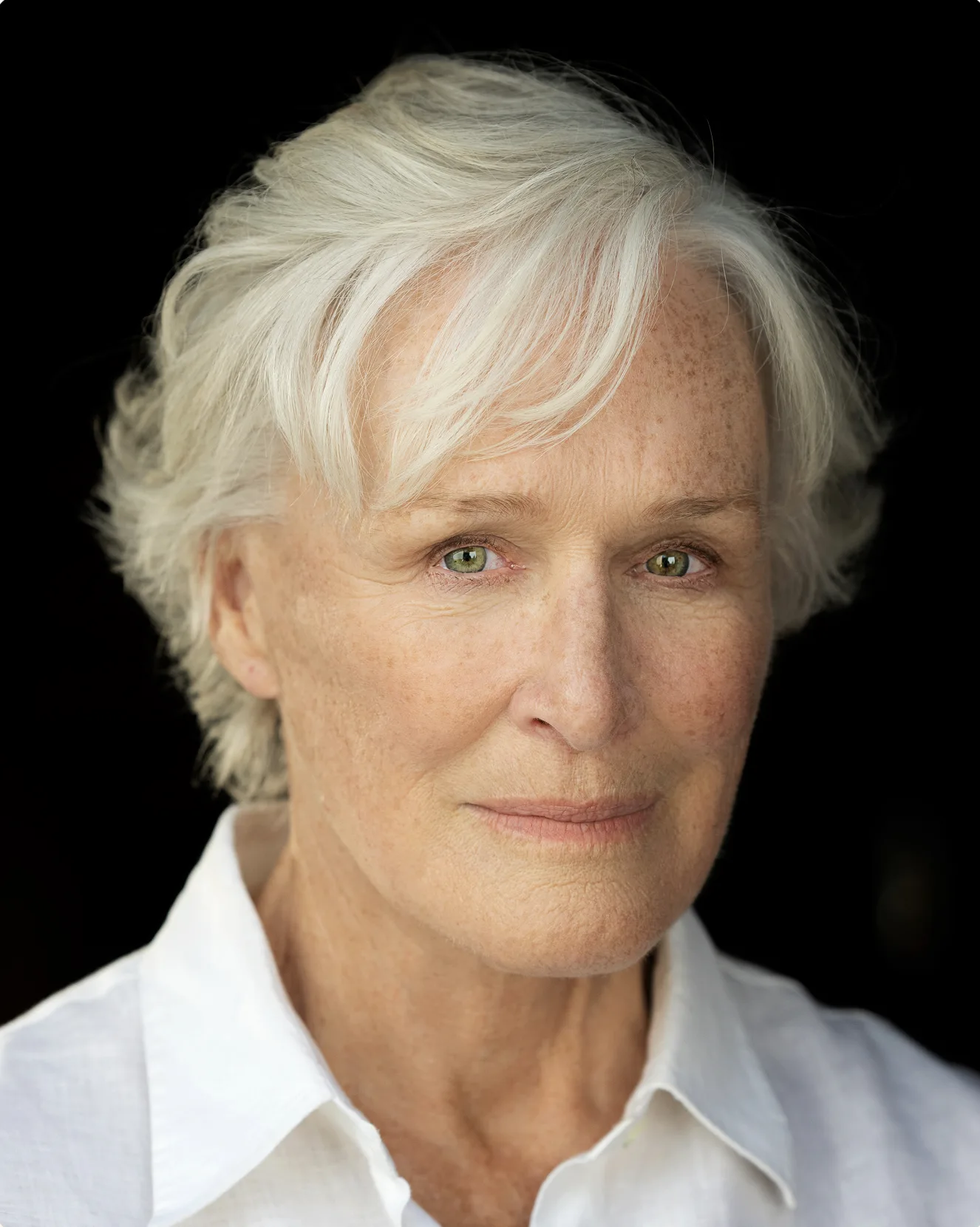 Head and shoulders portrait of Glenn Close with short white hair, wearing a white open-collared shirt against a black background