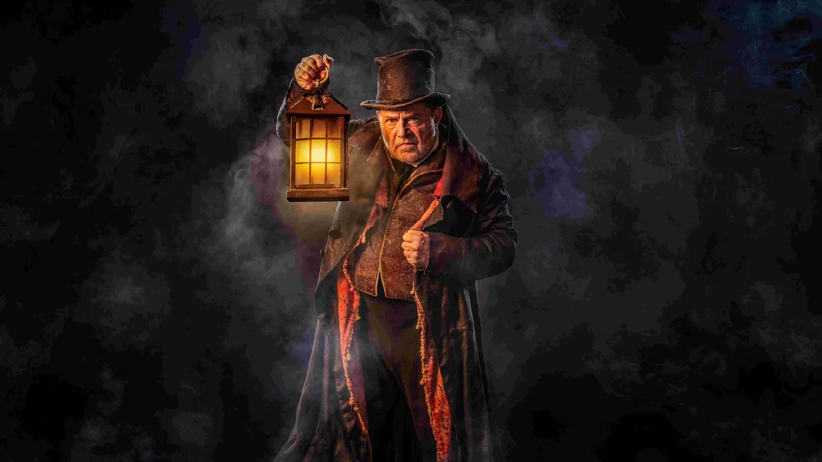 John Thomson as Ebenezer Scrooge holding a glowing lantern in Victorian costume for A Christmas Carol