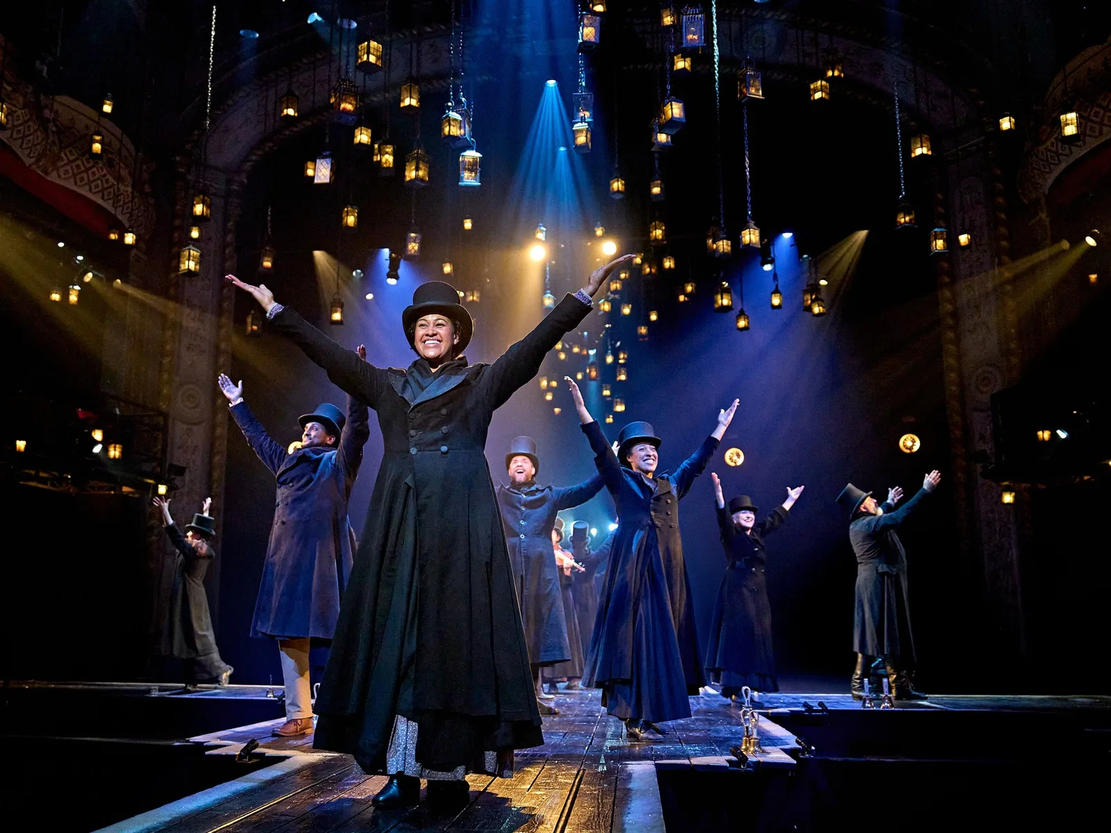 Company of A Christmas Carol on a lantern-lit stage in Victorian coats and top hats with arms raised