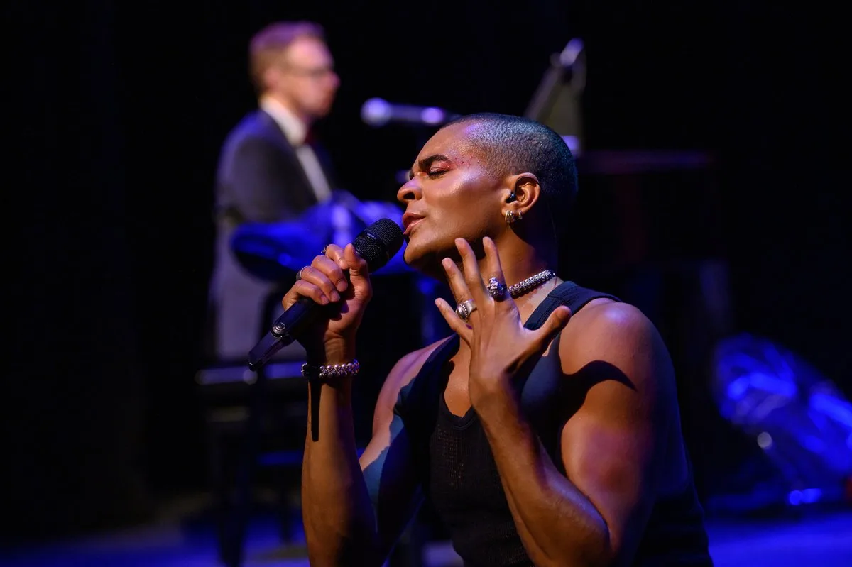Layton Williams sings into a microphone with his eyes closed, one hand raised beside his face, as a pianist plays behind him