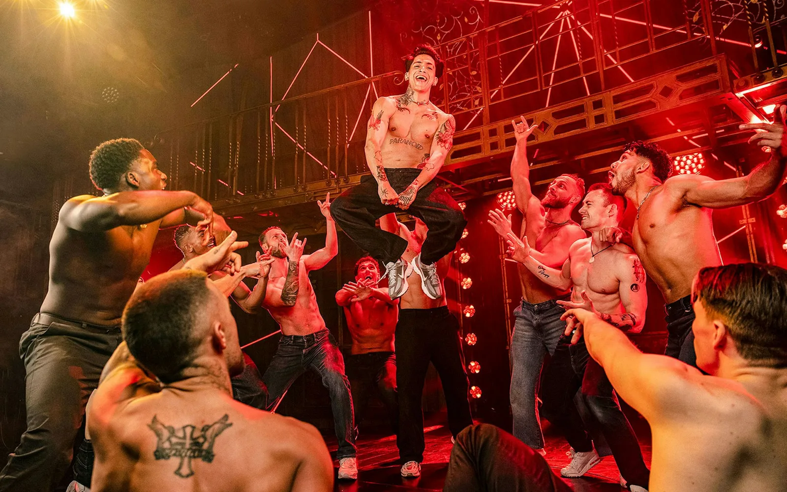 Magic Mike Live performers leaping and cheering on a red-lit stage with a metal gantry behind them