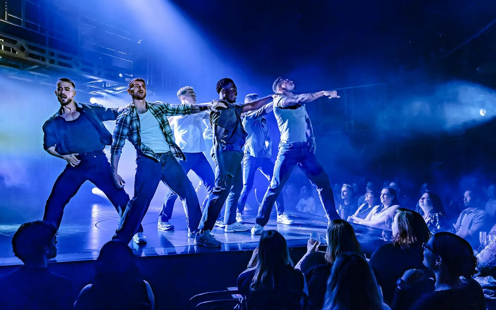 Six Magic Mike Live dancers in denim moving in formation under blue light as the audience watches from close by