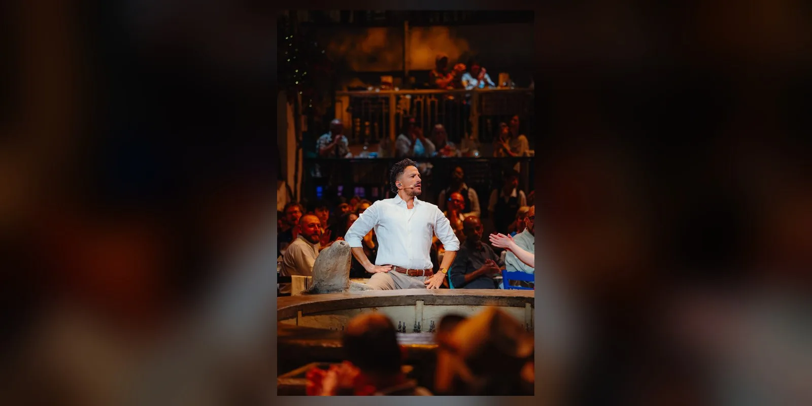Peter Andre in a white shirt with hands on hips as Nikos, with diners seated behind him