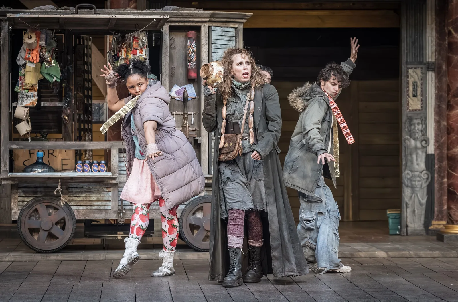 Three actors in ragged winter costumes stride across a wooden stage in front of a battered wagon hung with bric a brac