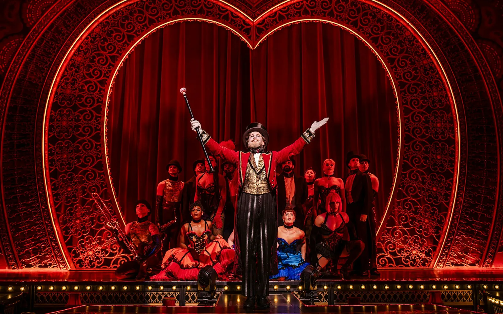A performer as nightclub owner Harold Zidler, in red tailcoat and top hat, raises his cane in front of a giant red heart